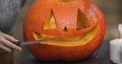 Person carving a pumpkin into a Jack-O'-Lantern on an outdoor wooden table with autumn leaves.